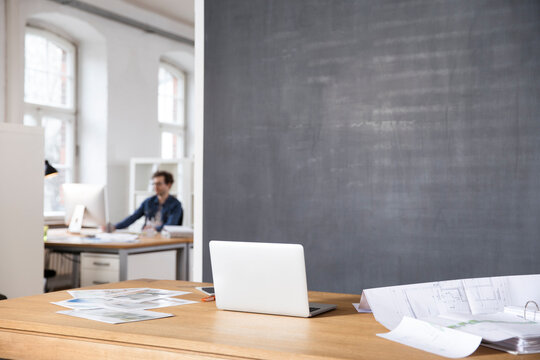Desk in office with laptop, photographies and blueprint and man in backgound