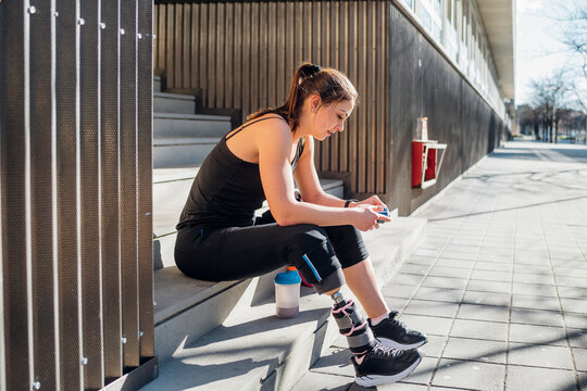 Sporty young woman with leg prosthesis sitting on stairs in the city using smartphone