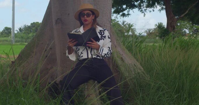 A peaceful scene in an expansive city park where a girl wearing sleek black trousers, a matching blazer, and a fashionable fedora enjoys a book