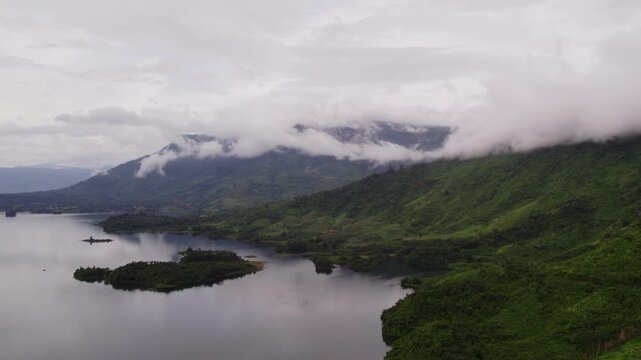 Sideward panning sliding airborne view of huge mountain surrounded by clouds and overcast sky, with large lake in the foreground.
