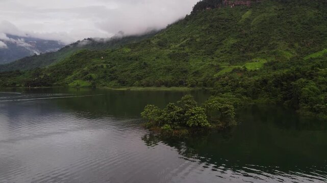 Circling rotaing flying view of small green island in the Nam Ngam Lake with green lush dense mountainous landscape in the background.