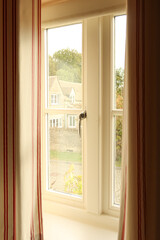 Cottage window with striped curtains, daylight shining through, offering a view of a traditional stone house and garden