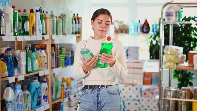 Young woman store visitor thinking about purchase, choose dish soap. Customer carefully reads label, doubts and makes purchase decision. Customer stares at assortment, spontaneous unplanned purchase. 