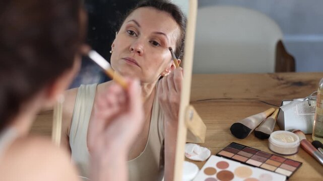 Focused woman carefully applying eyeshadow with brush while looking into vanity mirror, surrounded by makeup tools on table