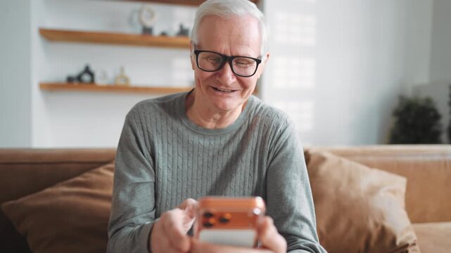 Phone addiction, aged man watching funny videos in social media, use smartphone. Portrait of laughing and smiling broadly old man resting alone in apartment, modern technology and new application