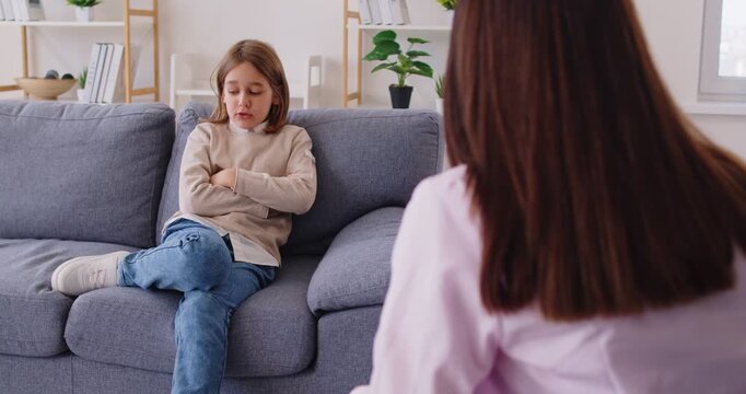 Frustrated little child sitting on sofa with arms crossed, avoiding eye contact with psychotherapist during counseling session. Child with signs of emotional distress talking to school psychologist.