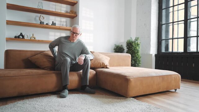 Aged male person suffering lower back pain, sitting on sofa in apartments. Disease of spine in old age, trauma or spinal injury and osteochondrosis, grey-haired man feeling ache and rubbing back