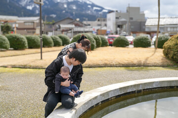 Fototapeta premium Father holds baby near water feature. Mother leans close, smiling gently. Background shows quiet town with mountains. Soft lighting enhances calm family moment. Ideal for parenting, family