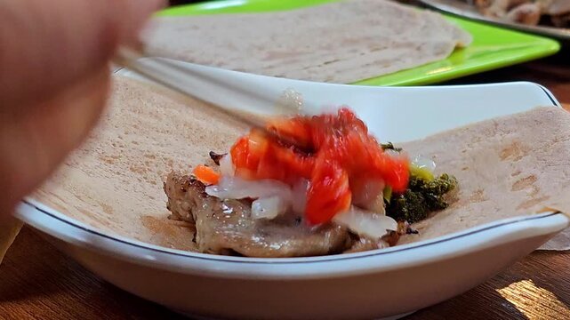 Extreme close-up of hands filling and rolling buckwheat crepe with boiled pork slices, kimchi, pickled daikon and carrot into tight bossam wrap in Korean restaurant