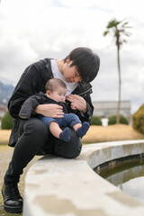 Young man holds baby gently, both smiling softly. They sit beside calm water, under soft cloudy sky. Palm tree stands tall in background, distant mountains loom. Moment captures quiet affection © Masakazu Tokashiki