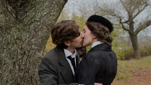 Vintage couple in historical clothing kiss near a tree in an autumn forest as the camera slowly moves around them in slow motion, creating a tender romantic moment in a peaceful natural landscape.