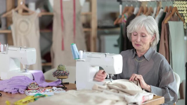 Older dressmaker works at a sewing machine against the background of racks with fabrics. Experienced clothing designer at work in a sewing workshop. High quality 4k footage