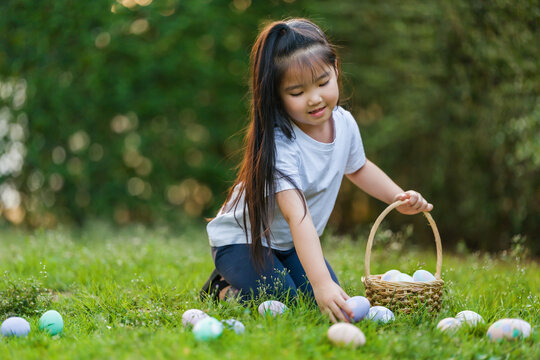 happy child girl playing and hunting for Easter eggs into a basket on green grass in garden