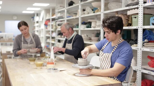 Young woman, young man and elderly man in ceramic workshop sculpting clay products during master class