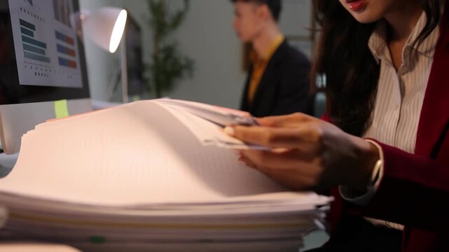 Businesswoman's hands sorting through a large stack of papers at her desk while working late at night, analyzing data charts on a computer.