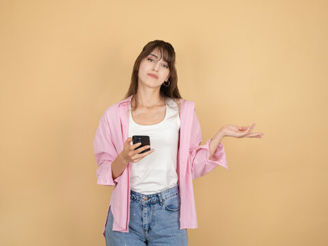 Young woman holding smartphone while raising open palm in uncertain gesture, wearing pink shirt and denim, standing against beige studio background wide copy space. Confusion and decision concept.