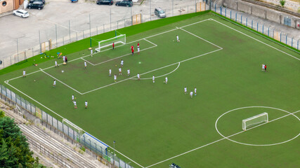 Obraz premium Aerial close-up of a vibrant green synthetic soccer pitch where two teams are engaged in a match. The field features white markings, two goals, and several players scattered across the turf.