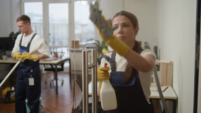 Focus on young Caucasian female cleaner spraying surface of glass door with detergent and wiping it while two colleagues working in background