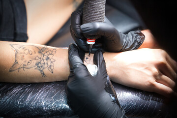 Fototapeta premium Detail of a female tattoo artist's hands, wearing black gloves and holding a tattoo machine, tattooing another woman's arm in her studio. Concept: ink, needle, tattoos, design, tending.