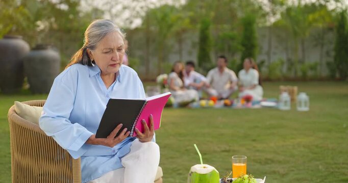 Indian old aged relax woman mom sitting on chair hold book read story novel outdoor house place happy male female man group girl child kid play spend quality time enjoy new day open in air garden lawn