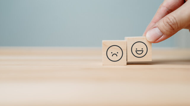 Hand placing wooden block with smiling face next to sad face cube representing customer satisfaction improvement, positive feedback, emotional change, and service quality evaluation concept.