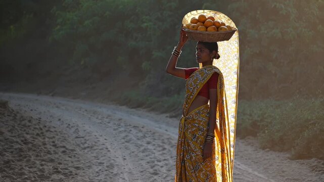 Woman vendor balancing basket of orange on head. Sari and bangle visible. Beach path and sunset light frame figure. Travel and tourism lifestyle theme. Cultural market portrait. Sunlight warm glow.