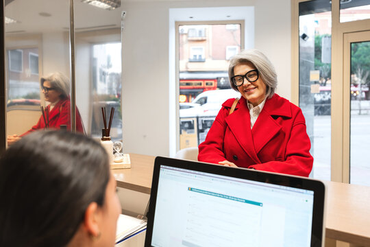 Smiling senior woman wearing a red coat at a dental clinic reception, checking in for an appointment with the staff