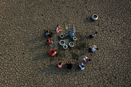 Aerial view of women and children gathered around pots on cracked earth, a stark contrast against the parched landscape, Kolkata, West Bengal, India.