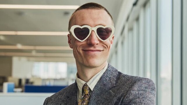 Confident mature businessman wearing playful heart shaped sunglasses posing in modern office interior smiling with ironic charm and unconventional executive attitude