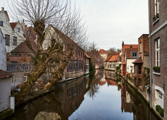 Obraz premium Serene canal in historic Belgian town Bruges. Medieval brick houses with gables reflect in calm water conveys concepts of heritage architecture, seasonal melancholy and urban waterway symmetry