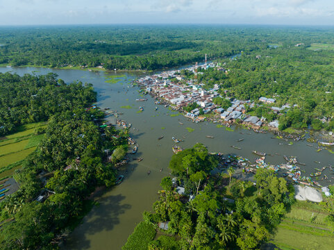 Aerial view of the river winding through lush green landscapes and a bustling riverfront village, homes lining the waterway, creating a vibrant scene, Barishal, Barisal Division, Bangladesh.