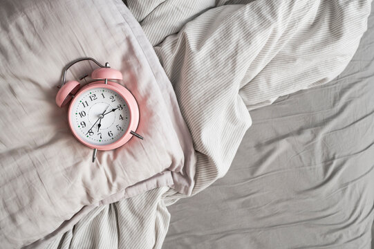 Pink alarm clock in bed on gray pillow and blanket, top view. Concepts of morning, waking up, healthy sleep or insomnia, circadian rhythms, daily routine

