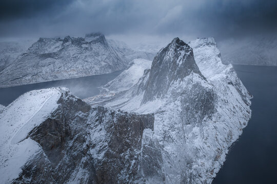 Aerial view of jagged, snow-dusted peaks rise majestically from dark waters under a brooding sky, the stark landscape a study in contrasts, Senjahopen, Troms, Norway.