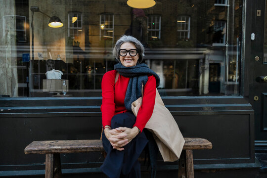Smiling shopper with scarf and eyeglasses sitting on bench outside storefront