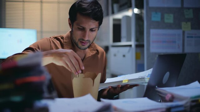 Overworked businessman facing deadline pressure while working late at night in the office, eating noodles a busy paperwork and nonstop work after hours