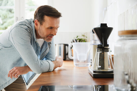 Man leans on kitchen counter watching drip coffee maker in morning