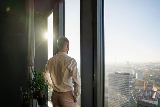 Young man in modern office looking out of a glass window at sunset