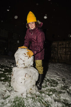 Teenager building a snowman in a snowy garden at night in winter