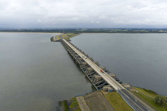 Aerial view of the Haringvlietdam stretching across the water, a concrete and steel behemoth under a cloudy sky, contrasting with the green shores, Haringvlietdam, South Holland, Netherlands.