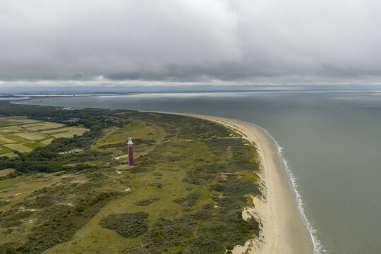 Aerial view of the Westhoofd Lighthouse standing tall amidst the green dunes and the sandy beach meeting the calm sea under a cloudy sky, Ouddorp, Zuid-Holland, Netherlands.
