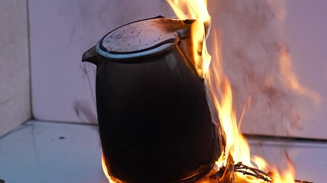 A electric kettle engulfed in flames, with the fire brightly illuminating its surface and rising upward. a dangerous situation, with dense tongues of fire covering its body.