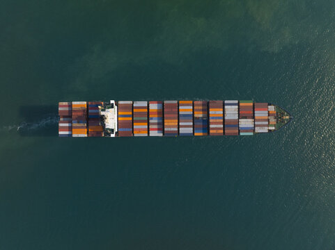 Aerial view of a long, fully loaded container ship cutting through the deep turquoise waters, leaving a frothy white wake behind, Pattaya City, Chon Buri, Thailand.