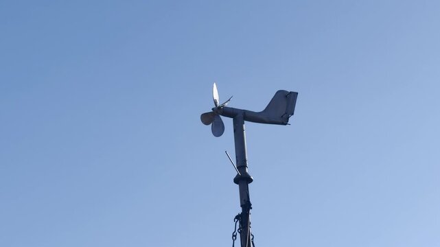 Weather station wind vane and anemometer rotating on a mast against a clear blue sky. Meteorological instrument measuring wind direction and speed, concept of climate monitoring and forecasting.