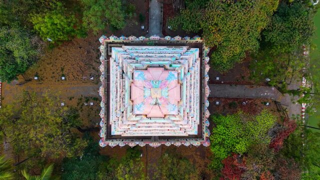 Aerial top-down view of a temple shrine centered on a square island within green water, surrounded by dense trees, palm canopy and pathways creating a striking geometric pattern in a historic tank.