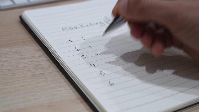 Close-up of a hand writing numbered steps for a marketing plan in a spiral notebook with a pen on a desk, representing business planning, strategy development, and idea organization in a workspace.