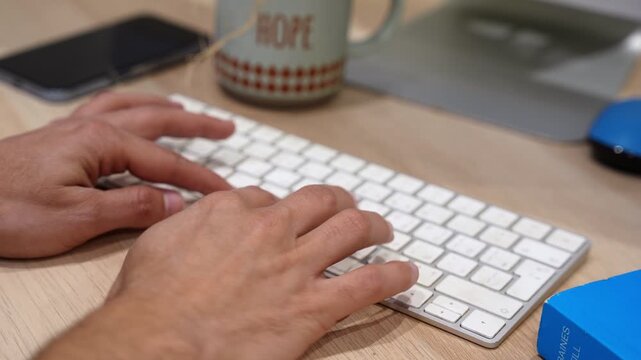 Close-up of hands typing on a computer keyboard at a wooden desk with a coffee mug, smartphone, and monitor nearby, representing productivity, office work, communication, and modern workspace activity