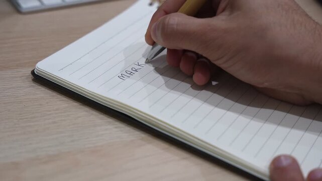 Close-up of a hand writing numbered steps under a marketing plan title in a spiral notebook on a desk, illustrating business planning, strategy development, and organized workflow ideas.
