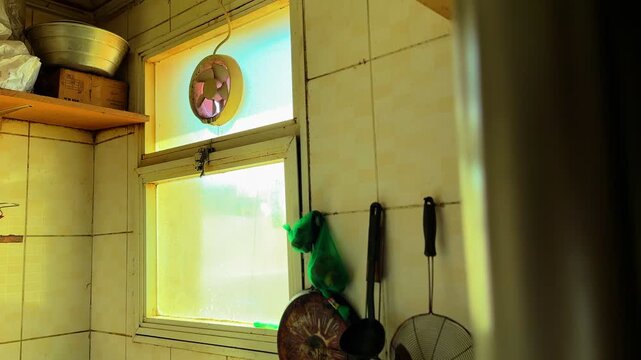 Warm sunlight entering through a frosted kitchen window with a small ventilation fan, rustic shelves, and cookware in a modest home interior, capturing everyday domestic life and natural indoor lighti