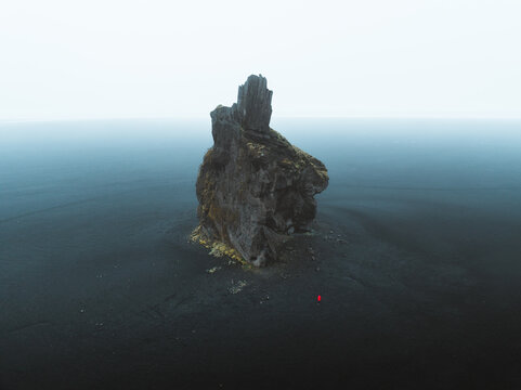Aerial view of Hjorleifshofdi standing stark against the dark sand, a solitary monolith amidst the Icelandic coast, Myrdalshreppur, Iceland.