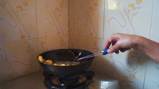 Closeup shot of Indian woman deep frying snacks at home on stove. Authentic process of preparing traditional Indian snack Chakri. Woman wearing bangles in hand putting snack into hot oil for frying.